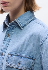 Svetlo modra denim srajca z ostrim ovratnikom, vidnimi šivi in prsnim žepom z gumbom ter majhnim logotipom.
