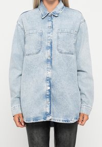 Žena v osvětlené modré oversized džínové košili s rozpuštěnými knoflíky a dvěma náprsními kapsami, doplněné o tmavé kalhoty.