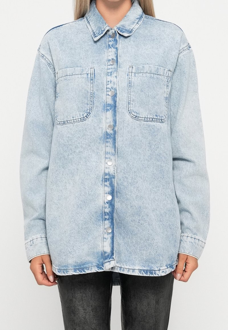 Žena v osvětlené modré oversized džínové košili s rozpuštěnými knoflíky a dvěma náprsními kapsami, doplněné o tmavé kalhoty.