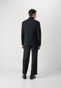 Giacca nera con schiena dritta e maniche lunghe, abbinata a pantaloni coordinati. Tessuto strutturato, dettagli di cucitura minimi e colore uniforme.