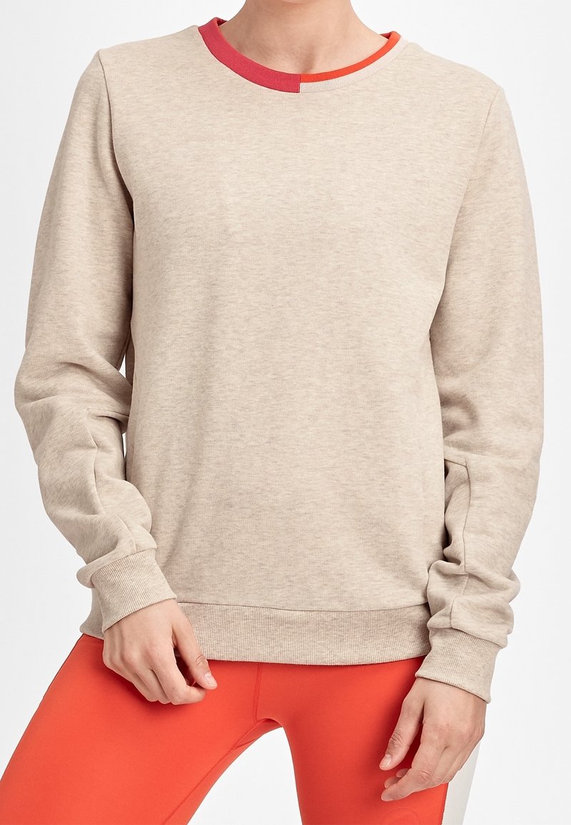 Personne portant un sweat-shirt à manches longues beige avec un détail de col rouge et des leggings orange vif, debout les mains près des hanches.