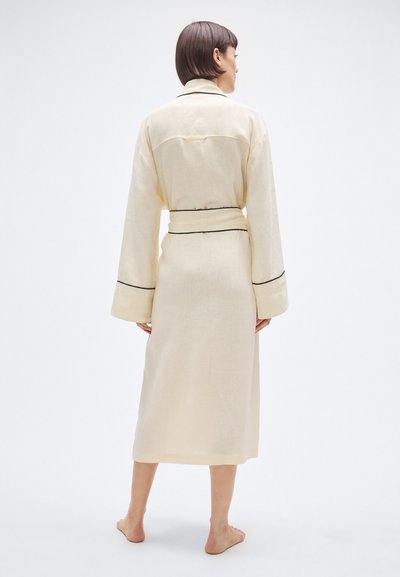 Bēšs lina halāts ar garām piedurknēm, jostu viduklī un melnu apmali aprocēs un apkaklī. Redzams no aizmugures.