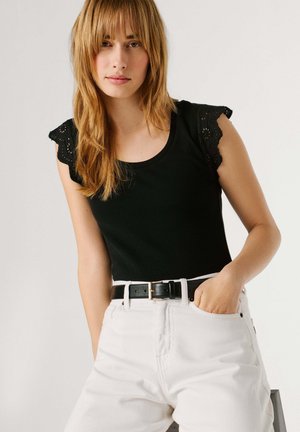 Kvinde med langt blondt hår iført sort ærmeløs top med blonder og hvide højtaljede jeans med sort bælte, hånd i lommen.