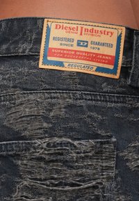 Noberzēti tumša džinsa bikses ar nolietotu ādas uzlīmi, uz kuras ir rakstīts "Diesel Industry Denim Division" un zīmola detaļas.
