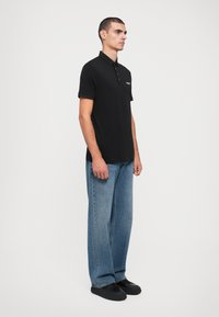 Svart polo shirt med korte ermer, tre-knappers placket og logo på brystet, kombinert med lyseblå vide denimjeans og svarte sko.