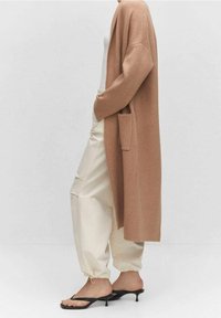 Larga chaqueta marrón de tejido suave, con apertura frontal y bolsillos laterales, combinada con pantalones crema holgados y sandalias negras de tacón.