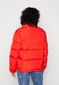 Doudoune rouge avec un col montant et des sections matelassées horizontales. Le tissu a une texture lisse. Portée par-dessus un jean bleu avec des accents brodés.