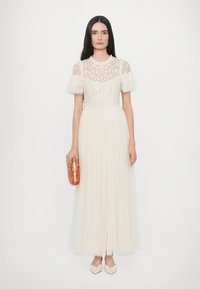 Lange, vloeiende jurk in crème met een doorschijnende, geborduurde bovenkant en geribbelde mouwen; heeft een textuur rok en een roze kralen clutch.