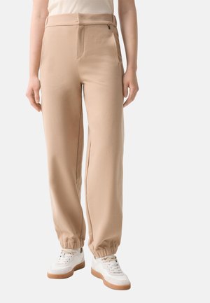 Beige Freizeithose mit elastischen Bündchen, getragen mit weißen Sneakers und weißem Oberteil, gezeigt von der Taille bis zu den Füßen.