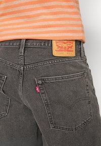 Personne portant un jean noir Levi’s avec une étiquette beige visible et une étiquette rouge, assorti d’une chemise à rayures orange et beige.