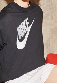 Musta puuvillainen t-paita, jossa on suuri valkoinen Nike-logo, pyöreä kaula-aukko ja lyhyt hiha, yhdistettynä valkoisiin shortseihin.