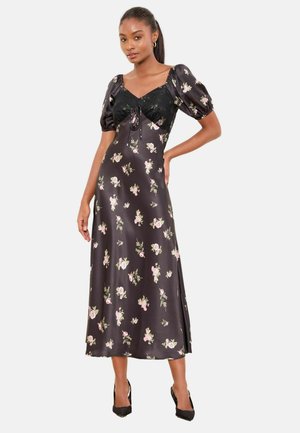 Frau trägt ein schwarzes Satin-Midikleid mit pinkfarbenem Blumenmuster und Puffärmeln, kombiniert mit schwarzen spitzen High Heels.