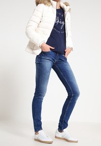 Vit puffjacka med fuskpälskrage, marinblå t-shirt med vit text, blå tajta jeans och vita sneakers med ljusgröna ränder.