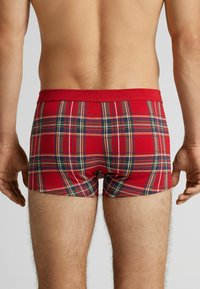 Czerwone bokserki w kratkę z czerwoną gumką w pasie, z zielonymi, żółtymi i niebieskimi liniami w wzorze. Tkanina wydaje się miękka i elastyczna.