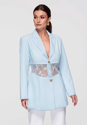 Vrouw draagt een lichtblauwe blazer met een kanten paneel bij de taille, gouden knopen, witte broek en gouden oorbellen in hartvorm, kijkt iets naar links.