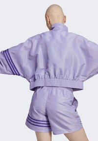 Vaaleanlila kevyt bomber-takki, jossa on joustava helma ja leveät hihat, yhdistettynä samansuuntaisiin shortseihin, joiden sivuilla on kaksi violettia raitaa.