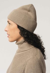 Beige gebreide muts met een ronde bovenkant, ribbelstructuur en een strakke pasvorm, gedragen met een bijpassende coltrui in een vergelijkbare kleur.