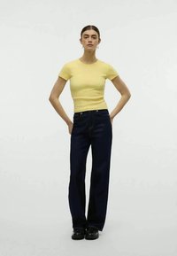 Top justo amarelo de mangas curtas combinado com jeans escuros de cintura alta. O modelo está de pé com as mãos nos bolsos, usando sapatos pretos.
