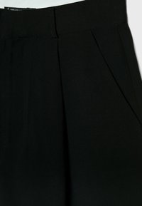 Pantalones negro ajustados con cintura entallada, que presentan pliegues frontales y una tela texturizada. Incluyen bolsillos laterales y detalles de costura limpia.