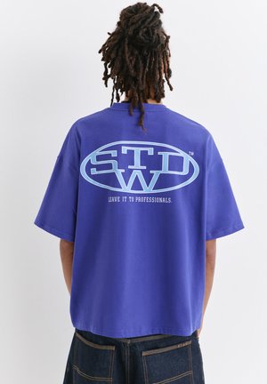 Homme avec des dreadlocks porte un T-shirt violet oversize avec le logo "STW" et le texte "Leave it to professionals." au dos, associé à un jean foncé.
