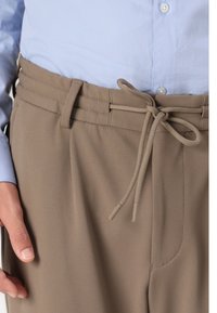 Beige teksturerte bukser med strikket midjebånd, vist sammen med en lyseblå skjorte med knapper. Nærbilde med fokus på midjen og stoffet.