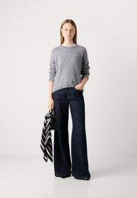 Mulher de pé, vestindo um suéter cinza e jeans largos escuros, segurando uma bolsa preta com um lenço listrado em preto e branco.