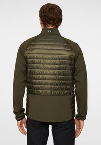 Henkilö, joka on pukeutunut vihreänoliivinväriseen vetoketjulliseen takkiin, jossa on pehmustettu selkäosa, nähtynä takaa yksinkertaista taustaa vasten.