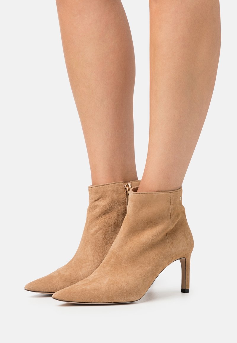 Femme portant des bottines en daim beige à bouts pointus et talons moyens fins, debout sur une surface blanche.