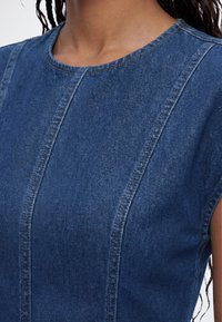 Κοντινό πλάνο ενός ατόμου που φοράει μπλε denim αμάνικο τοπ με κάθετες ραφές και στρογγυλή λαιμόκοψη.