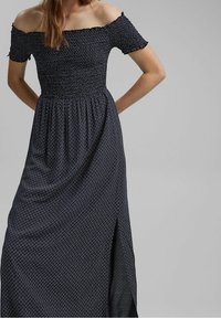 Abito maxi blu navy con spalle scoperte e corpino smock, caratterizzato da un motivo geometrico bianco e una fessura laterale per facilitare i movimenti.
