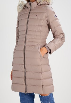 Femme portant un manteau matelassé beige mi-long avec capuche bordée de fourrure, fermeture éclair et main dans la poche, associé à un jean bleu déchiré.
