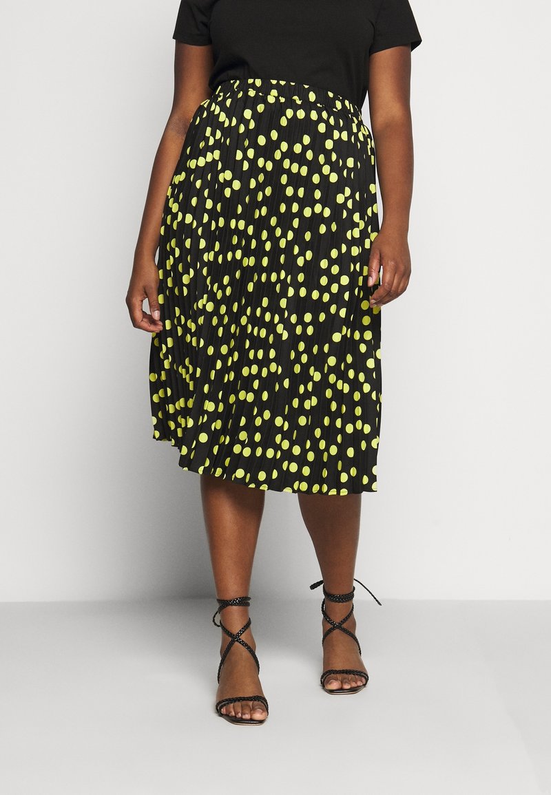 Falda negra con pliegues y grandes lunares amarillos, con una cintura elástica. Combinada con una camiseta negra y sandalias negras con tiras.