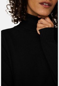 Frau mit lockigem Haar, die einen schwarzen Rollkragenpullover trägt und ihr Kinn an ihrer Hand neben ihren Lippen abstützt.