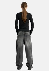 Kobieta z długimi brązowymi włosami w niskim, skręconym kucyku, ubrana w dopasowaną czarną bluzkę z długim rękawem i luźne, wyblakłe czarne spodnie typu wide-leg, stoi z rękami w kieszeniach.