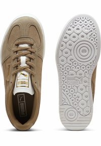 Beige mockask sneakers med rund vit sula. Har bruna snören, gulddetaljer på tungan och en strukturerad gummisula med cirkulära mönster.