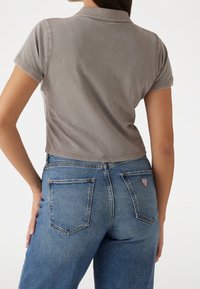 Kvinna som bär en grå kortärmad polo och högmidjade blå jeans, sedd bakifrån, med handen vilande på höfterna.