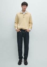 Maglione a maglia giallo chiaro con colletto e tre bottoni, abbinato a jeans di denim scuro e scarpe stringate nere. Il modello sta in piedi con le mani nelle tasche.