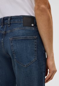 Granatowe dżinsy z bliska ukazujące detal tylnej kieszeni oraz czarną skórzaną naszywkę z białym emblematem krzyża.