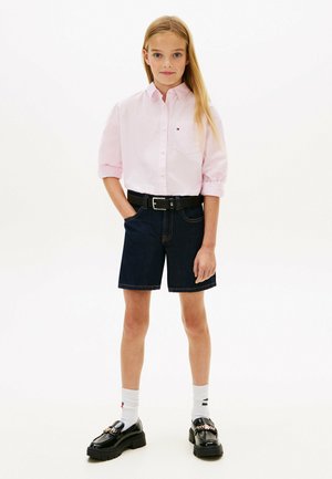 Jong meisje staat met handen in de zakken, draagt een lichtroze blouse met knopen, donkere denim shorts, een zwarte riem, witte sokken en zwarte loafers.