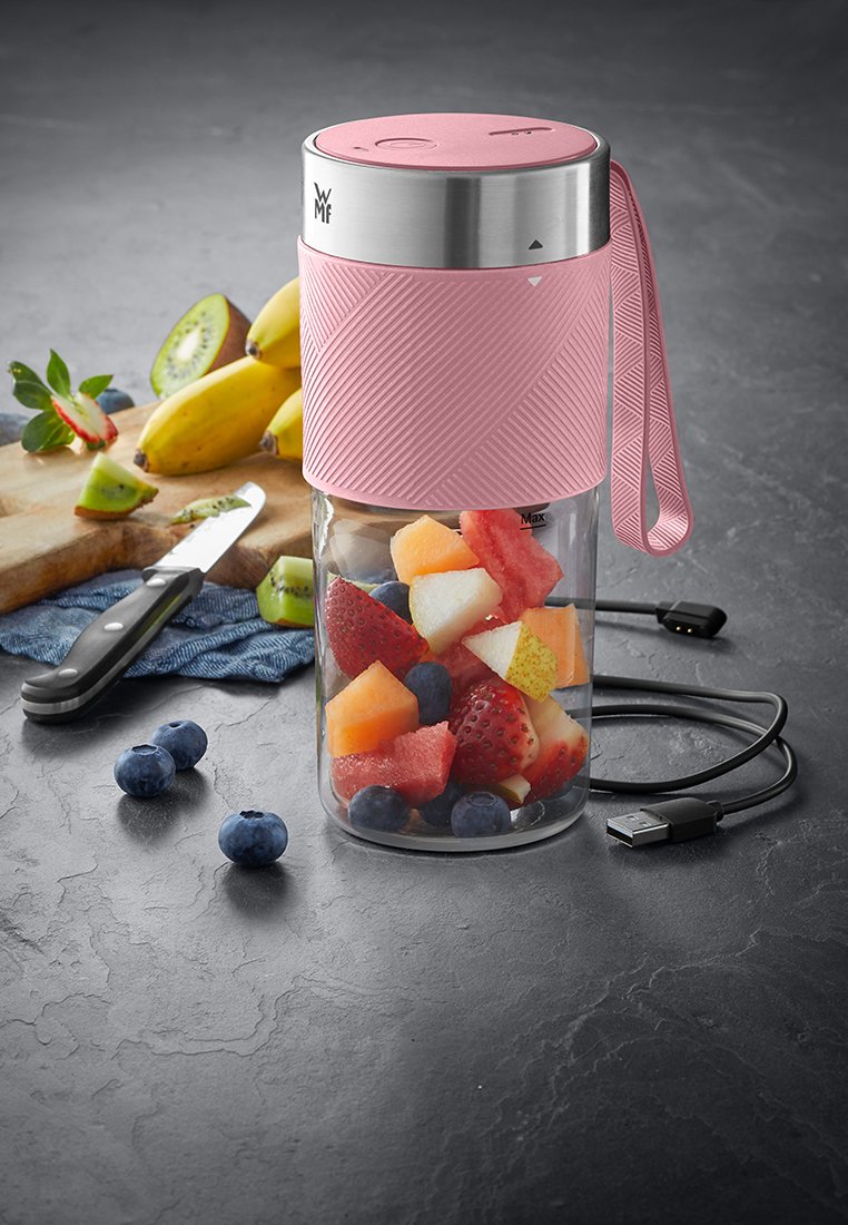Tragbarer pinker Mixer mit USB-Kabel, gefüllt mit gemischten Obststücken auf einer dunklen Arbeitsplatte neben einem Messer und Bananen.