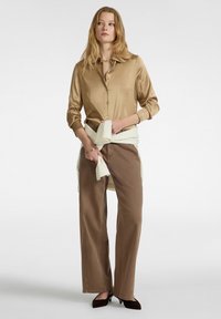 Ein satinierter, tannfarbener Button-Up-Hemd, kombiniert mit braunen, weit geschnittenen Hosen. Ein cremefarbener Pullover ist um die Taille gebunden. Schwarze Absätze vervollständigen das Outfit.