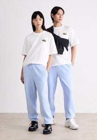 Camisetas de algodón blancas con logo bordado en verde, combinadas con sudaderas de color azul claro. Tejido texturizado y corte relajado, con zapatillas.