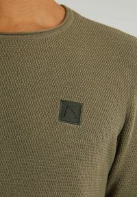 Olivenfarbener, strukturierten Pullover mit einem Wabenmuster, Rundhalsausschnitt und einem quadratischen Stoffpatch mit einem schwarzen Design auf der linken Brust.