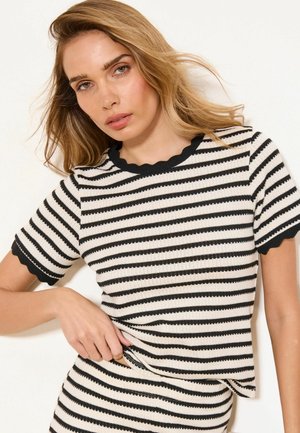 Kvinde med blondt hår iført en sort- og hvid horisontalt striped kortærmet top med bølget kant, der holder i kanten af toppen.