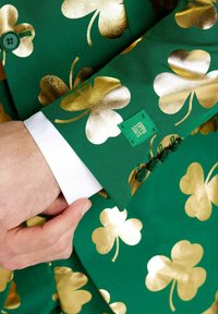 Zeleni blejzer s zlatnim vzorcem deteljice, z zelenimi gumbi in belim manšetnim delom srajce. Oznaka pravi "OPPO SUITS." Teksturovana tkanina.