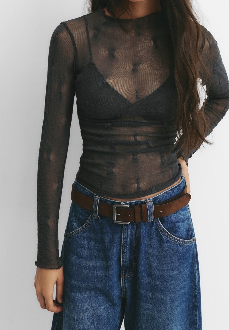 Femme portant un haut noir transparent à manches longues par-dessus un soutien-gorge noir, avec un jean bleu et une ceinture marron, debout devant un fond uni.