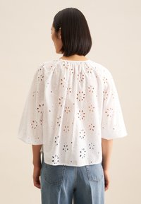 Blusa bianca con motivi in pizzo ricamati, scollo arricciato e maniche ampie. Abbinata a jeans in denim per un look rilassato.