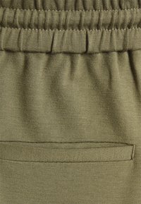 Tecido verde-oliva com uma textura suave e um cós elástico; apresenta um bolso plano com bordas cosidas na parte de trás.