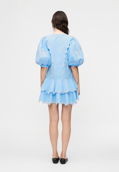 Γυναίκα που φοράει ένα ανοιχτό μπλε μίνι φόρεμα με φουσκωτά μανίκια και λεπτομέρειες δαντέλας με κυματιστό σχέδιο, στέκεται με την πλάτη προς τα εμάς, φορώντας μαύρα παπούτσια χωρίς κορδόνια.