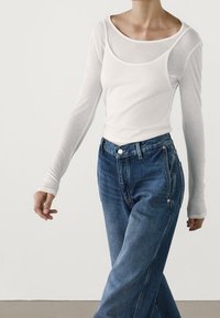 Top bianco a maniche lunghe sovrapposto a una canotta aderente bianca, abbinato a jeans in denim a vita alta di blu medio, con bottoni e tasche.